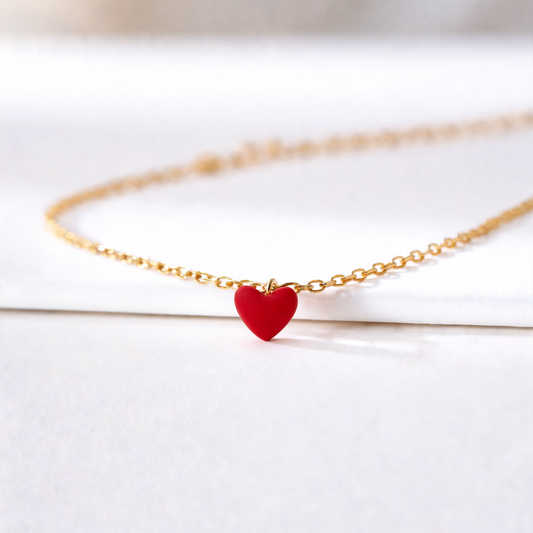 A delicate gold chain necklace featuring a small, glossy red heart-shaped pendant, displayed on a clean white surface with a soft-focus background.