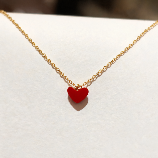 A delicate gold chain necklace featuring a small, smooth red heart-shaped pendant, displayed on a white surface.
