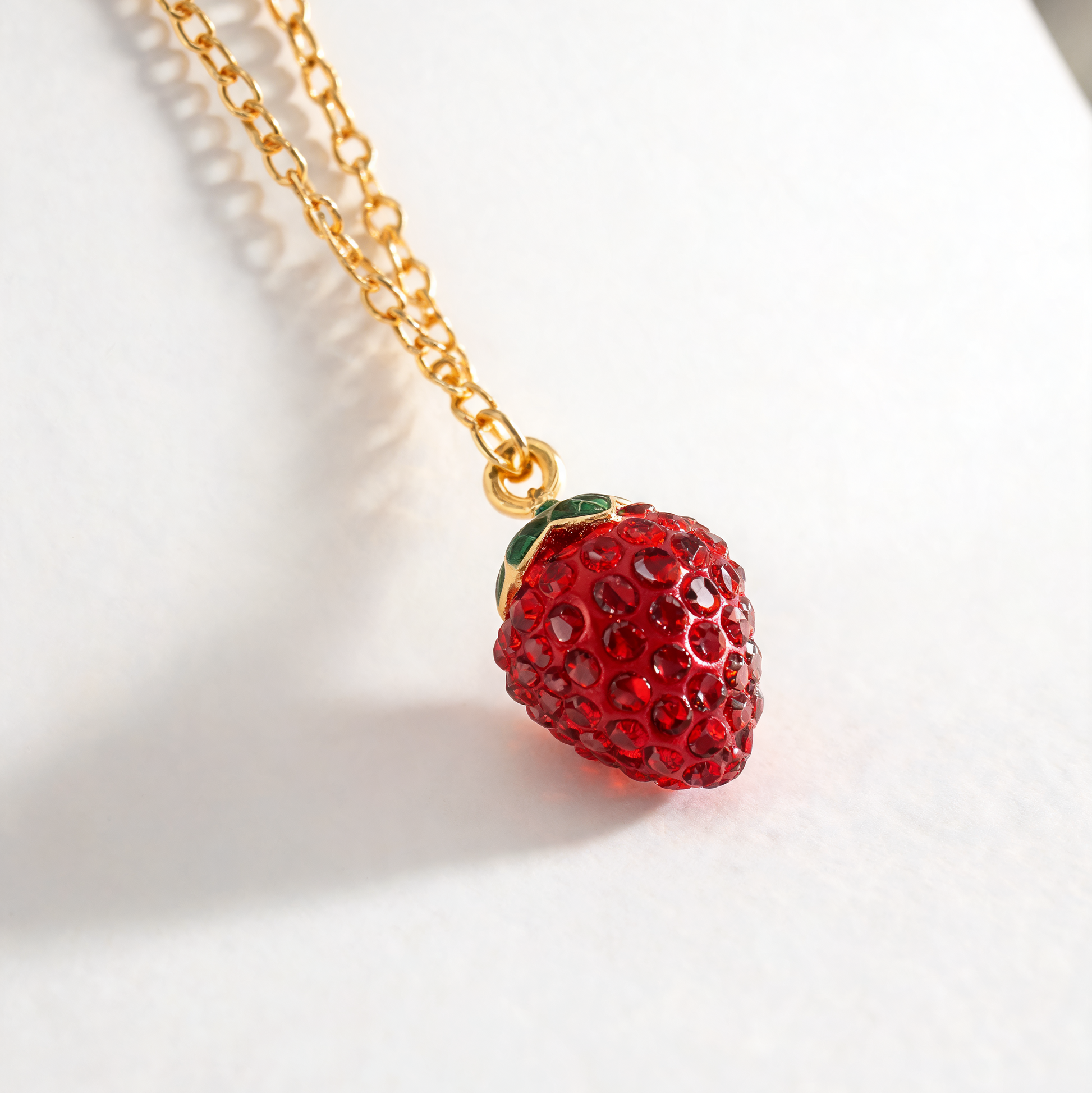 A gold chain necklace featuring a strawberry-shaped pendant encrusted with red rhinestones and accented with green enamel leaves, displayed on a clean white surface.