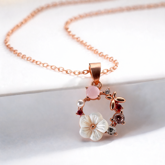 A rose gold chain necklace with a delicate wreath pendant featuring a white mother-of-pearl flower, a pink stone, and small accent stones, displayed on a white surface.