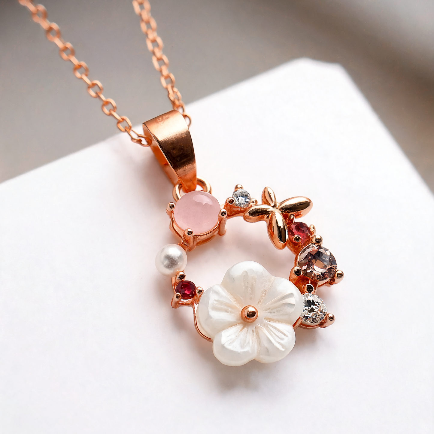 Rose gold floral pendant necklace with a white flower, pink stone, and small clear and red gemstones on a white background.