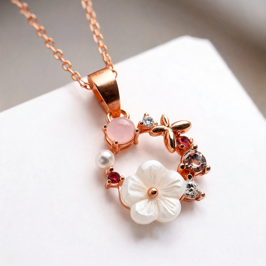 Rose gold floral pendant necklace with a white flower, pink stone, and small clear and red gemstones on a white background.
