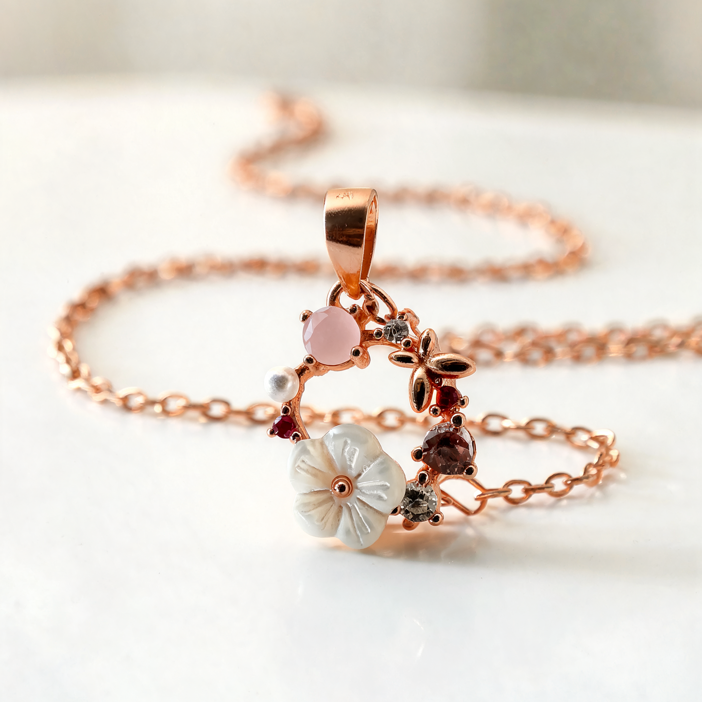 A rose gold chain necklace featuring a delicate pendant with a white flower, a pink flower, and small dark and silver gemstones, displayed on a soft white surface.