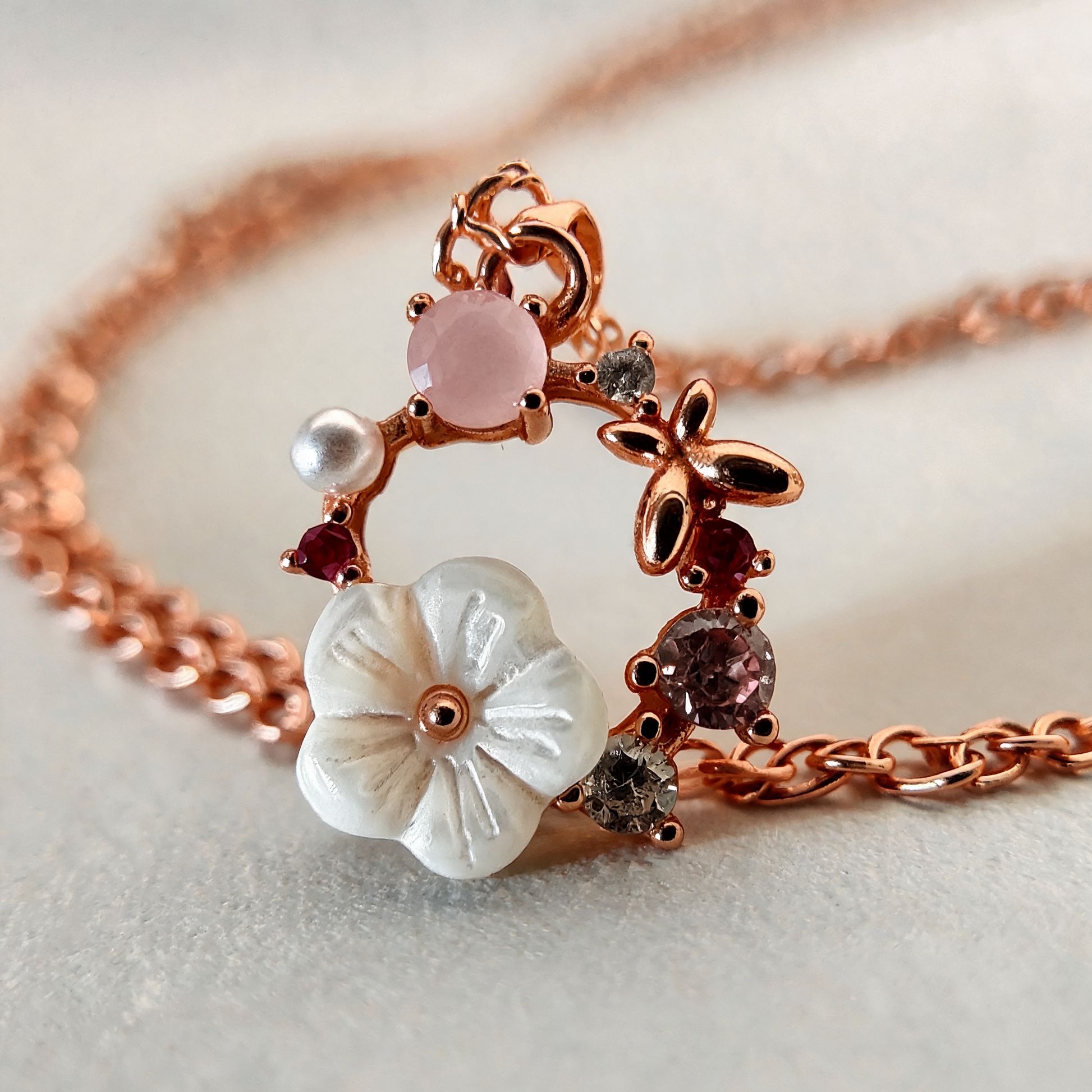 A rose gold chain necklace featuring a delicate floral and butterfly pendant. The pendant includes a large white carved flower, a pink cabochon stone, a small white pearl, and dark and grey gemstones, set against a soft neutral background.