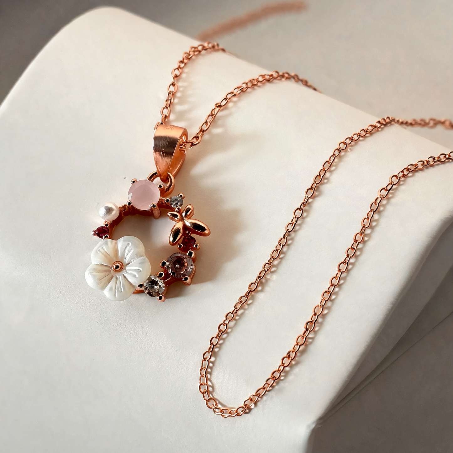 A rose gold chain necklace with a delicate floral pendant, displayed on a white jewelry bust. The pendant features a white flower, a pink flower, and small dark red and clear gemstone details.