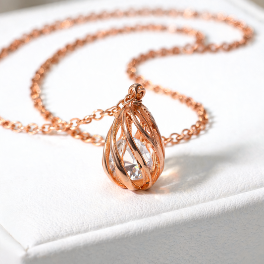 Rose gold necklace with a teardrop-shaped pendant featuring a twisted, flame-like design and a clear central gemstone, resting on a white textured jewelry box.