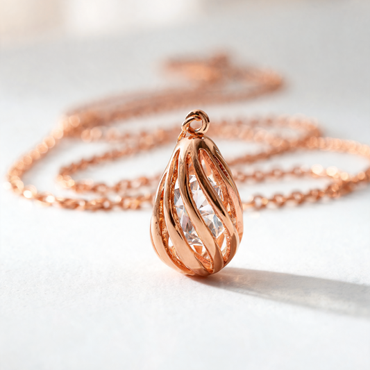 A rose gold teardrop pendant necklace with a textured, twisted design and a clear faceted center stone, resting on a light gray surface with its matching chain blurred in the background.