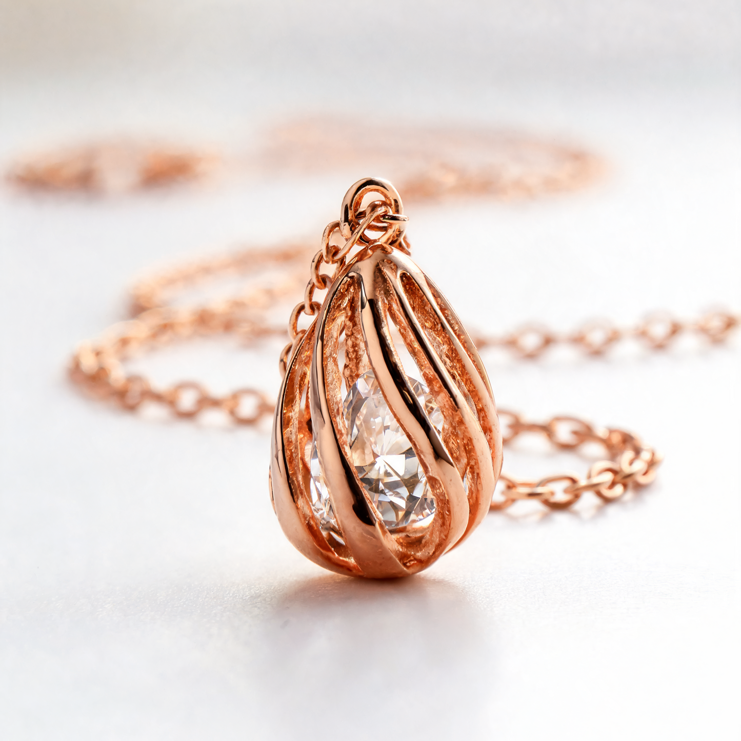 Rose gold teardrop pendant necklace with a clear faceted gemstone, shown on a soft white background with a blurred chain.