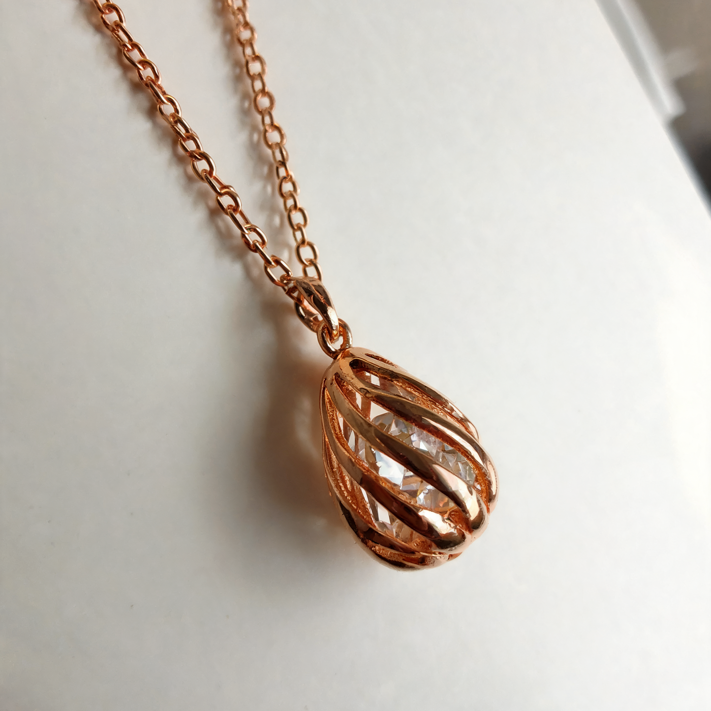 Rose gold chain necklace with a twisted, openwork teardrop pendant on a white background.